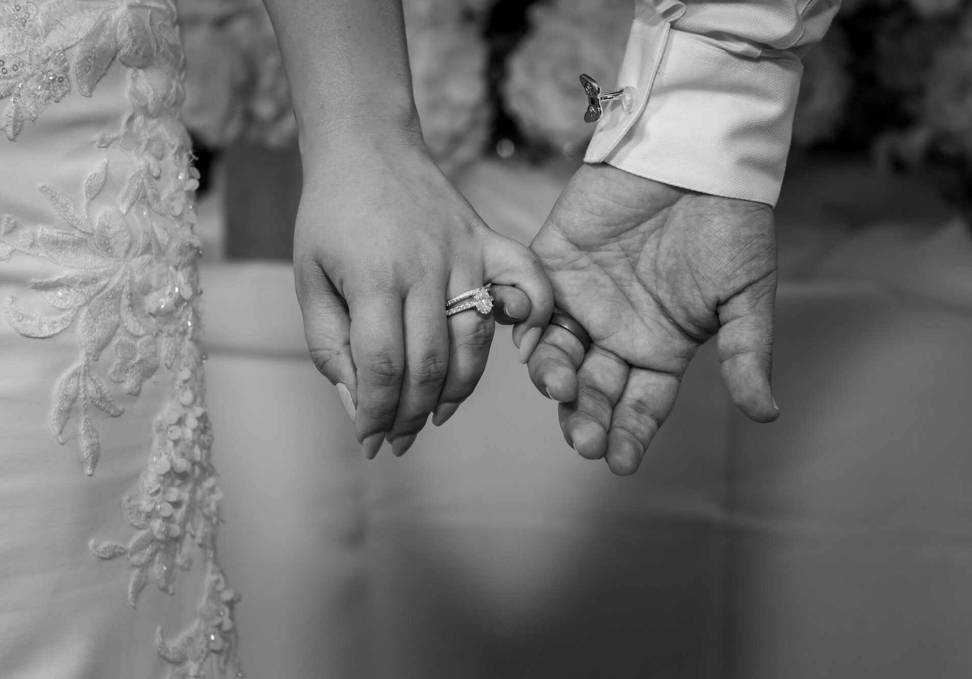 Black and white close-up of holding hands, showing their wedding rings and the lace details of her wedding dress.